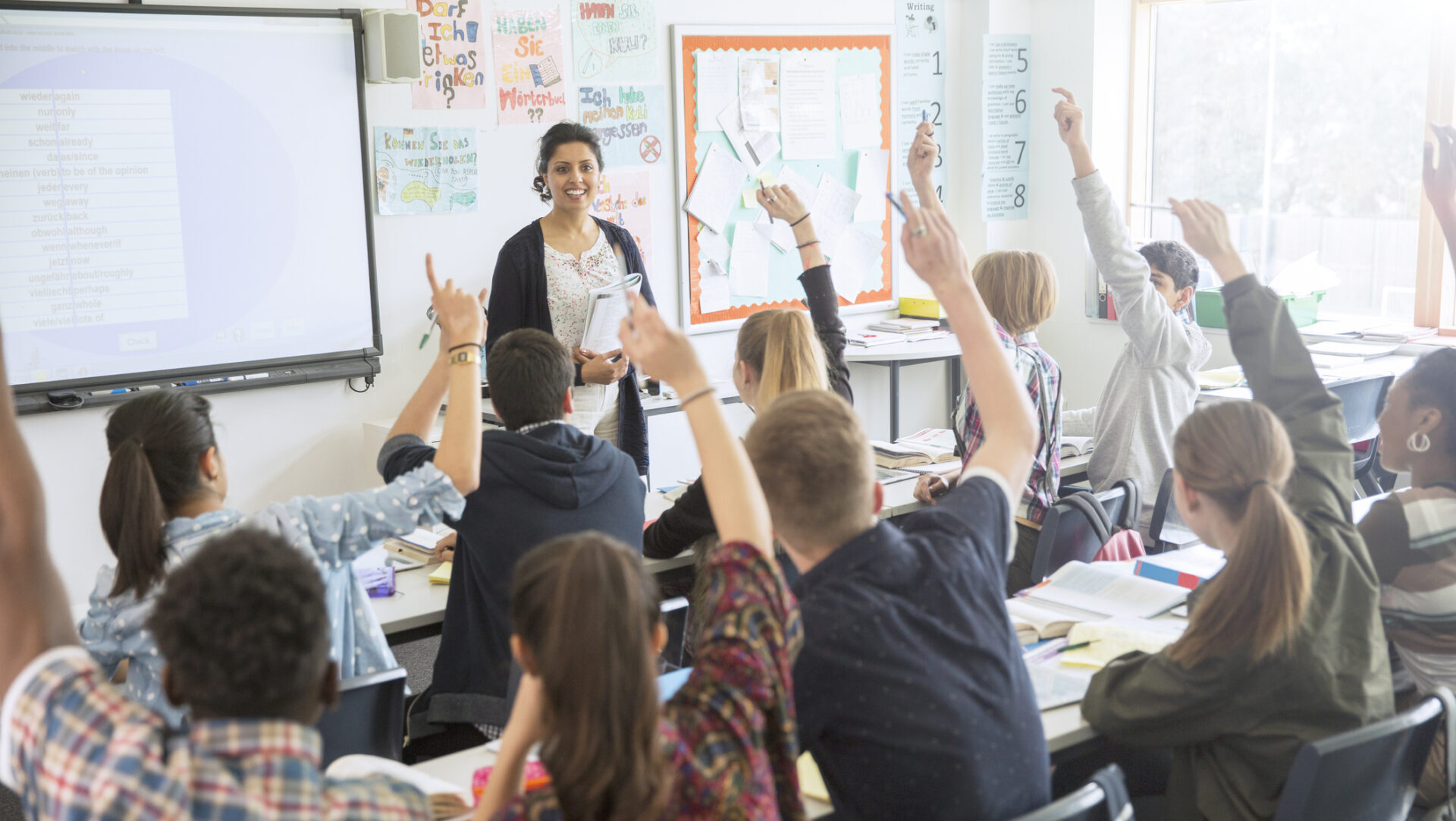 Rear view of teenage students raising hands in classroom Champion early financial education in your community.