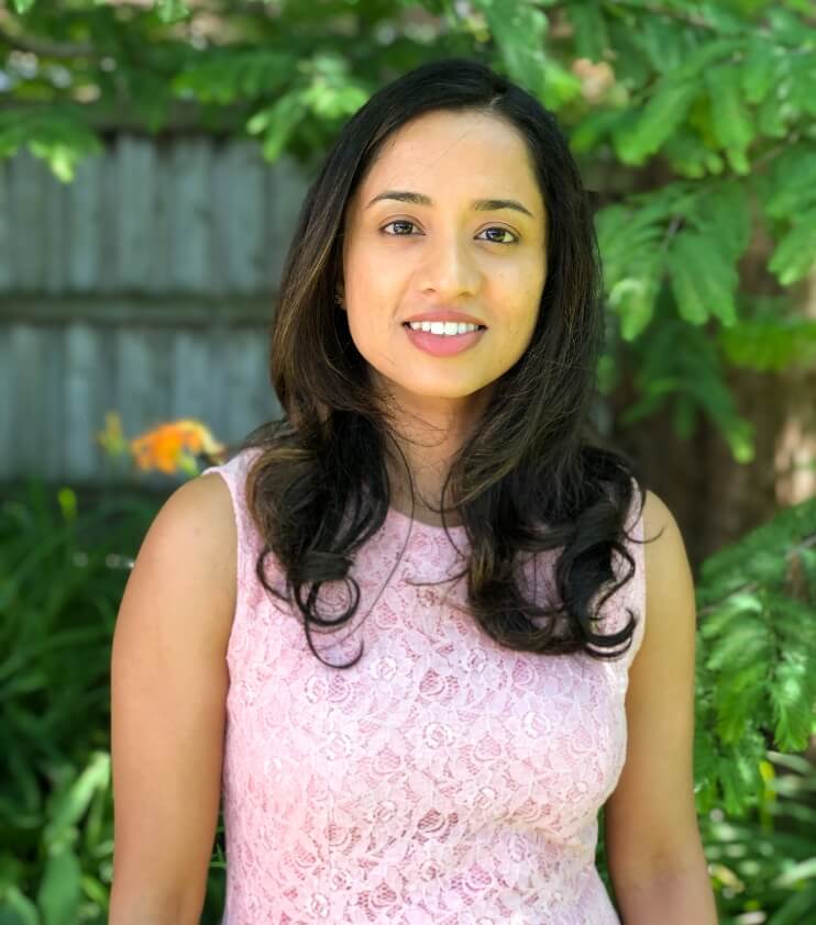 A woman with long dark hair wearing a light pink lace sleeveless top, standing outdoors with greenery in the background.