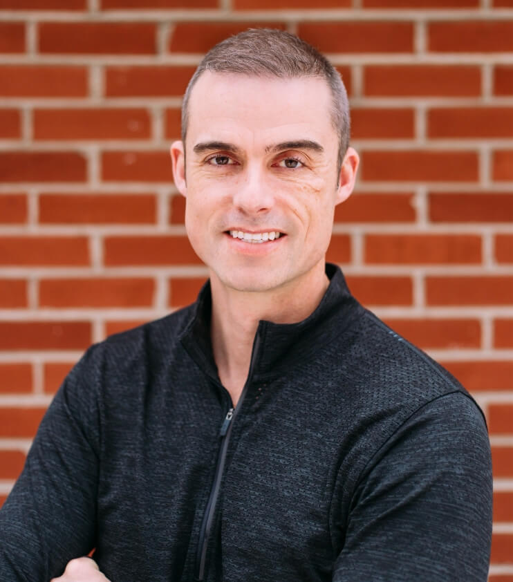 A smiling man with short-cropped hair wearing a dark quarter-zip sweater, standing in front of a red brick wall.